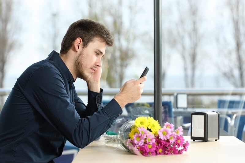 Sad man with a bouquet of flowers stood up in a date checking phone