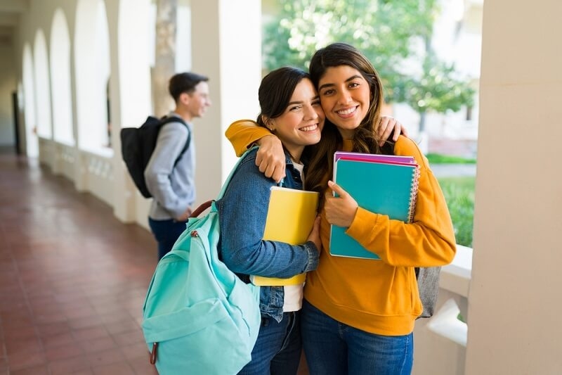 best friends with backpacks and books hugging and smiling in school