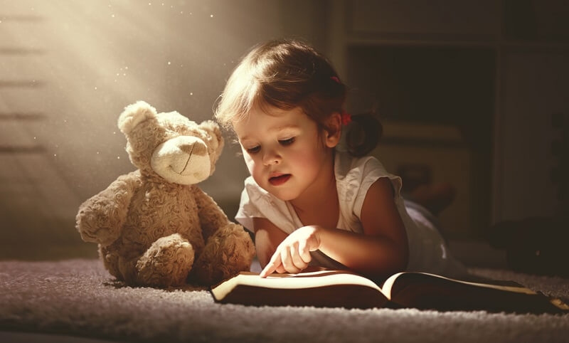 little girl reading a book with a toy teddy bear