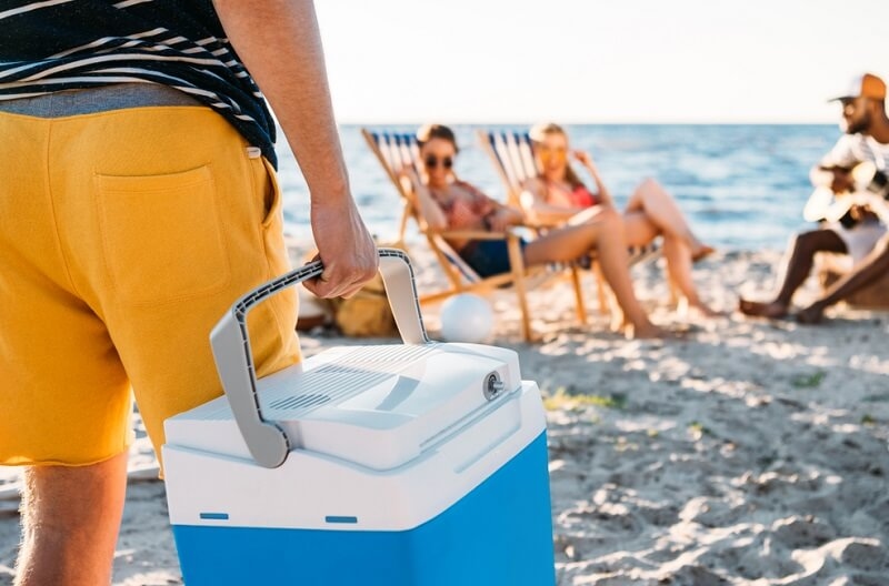 man carrying beach cooler while friends resting on sand behind