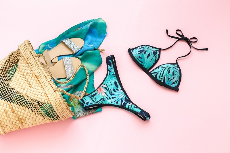Bikini swimsuit with tropical print, silver glitter flat sandans, wicker beach bag, sarong on pink background
