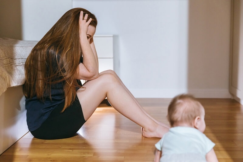 woman suffering from parental burnout putting hand on head and baby is sitting on floor
