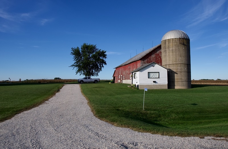 a long gravel driveway leads to a red barn, silo, and white outer building on a farm with a bright blue sky and a single tree in the background