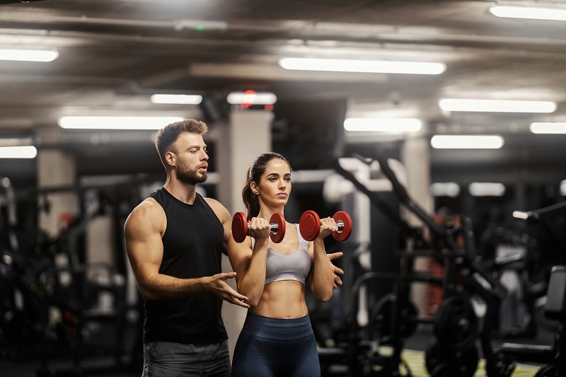 A fit sportswoman in shape is doing exercises with dumbbells in a gym and her coach is helping her.