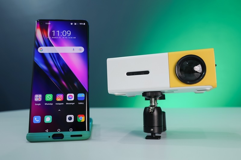 A mini projector on a table with a smart phone in a neon green background.