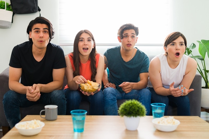 Surprised college friends watching a thriller movie or tv series on the sofa and feeling shocked while eating chips