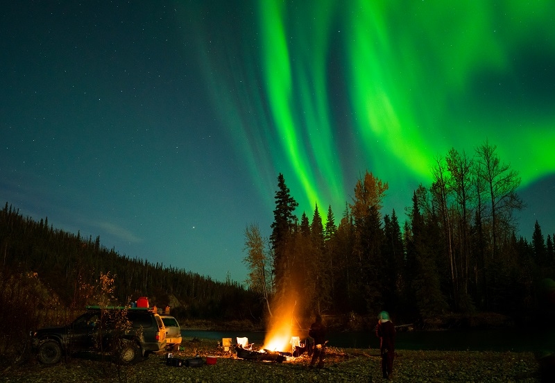 Northern Lights above people camping next to river with campfire
