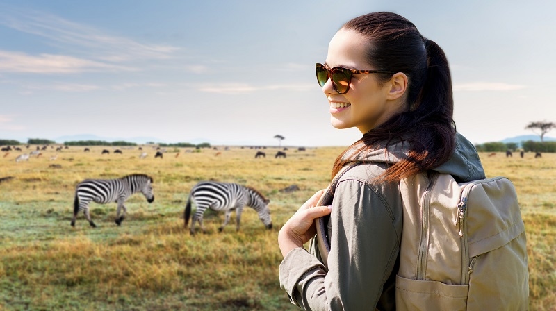 travel, tourism and people concept - happy young woman with backpack over african savannah and zebras background