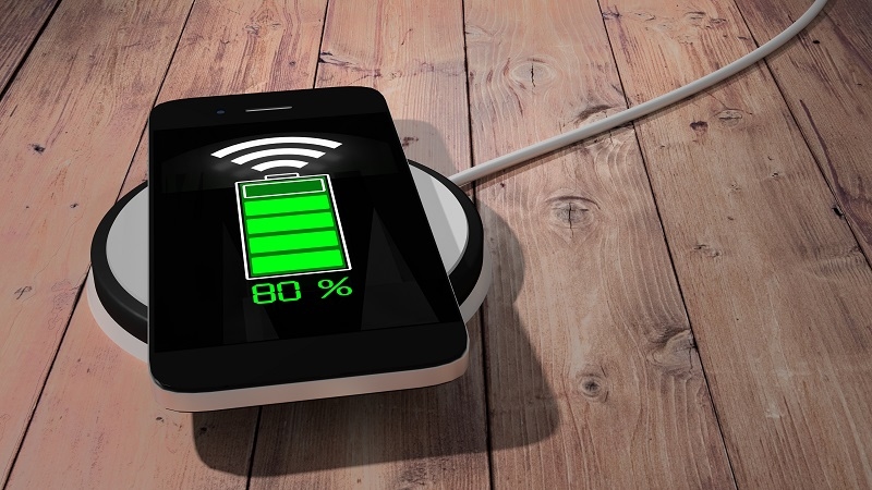 Wireless charging pad with mobile phone green glowing battery screen on wooden table 3D illustration