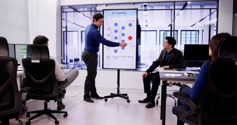 Team in a modern office attending an AI training session, with a presenter explaining a neural network diagram on a board.