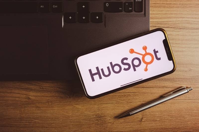 Smartphone displaying the HubSpot logo on a desk beside a laptop and pen, representing CRM and marketing software.