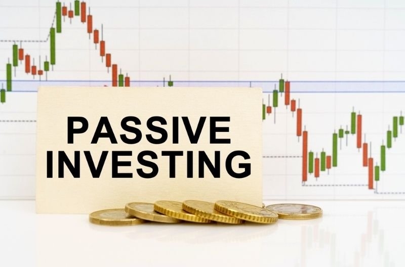 Sign reading “Passive Investing” placed in front of coins and a stock market chart, illustrating long-term investment strategies.