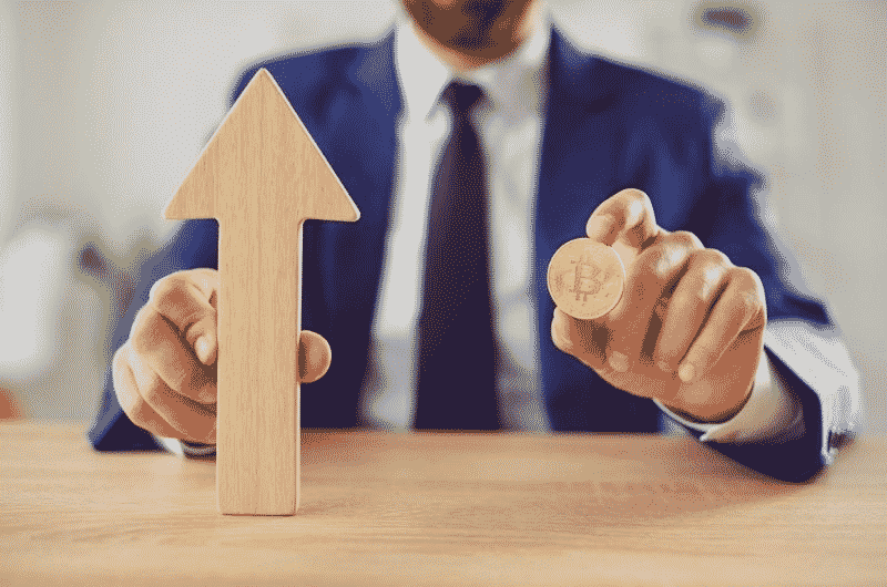 Businessman holding a Bitcoin coin next to an upward-pointing arrow, symbolizing cryptocurrency growth, investment, or rising market trends.