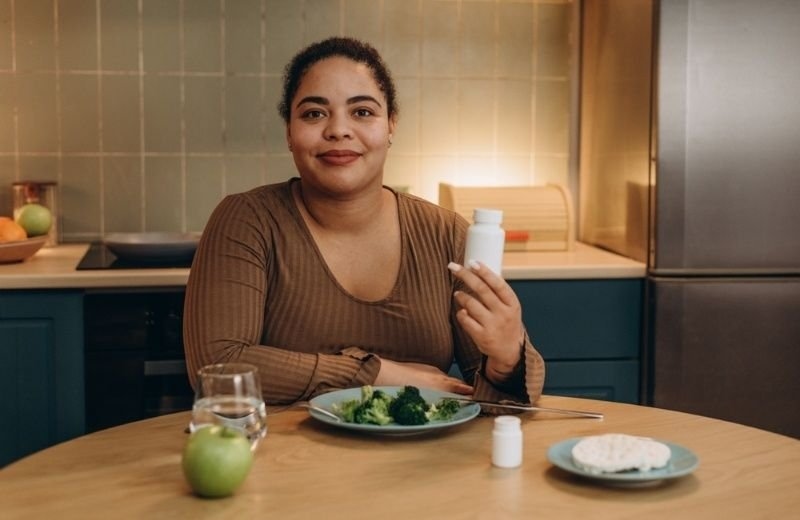 Woman sitting at a dining table holding a supplement bottle beside healthy food, representing wellness, nutrition, or postpartum recovery.