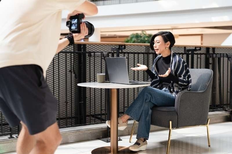 Woman recording a professional video or interview while sitting with a laptop as a camera operator films her.