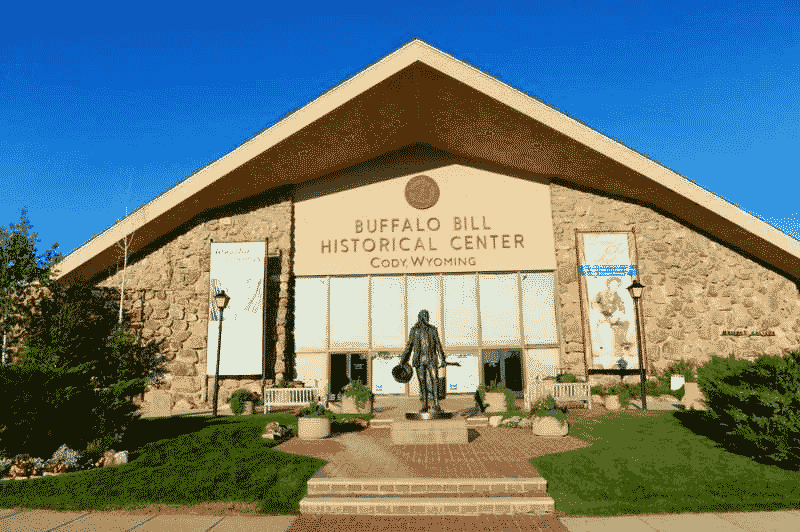 Front entrance of the Buffalo Bill Historical Center in Cody, Wyoming, featuring a stone building, statue, and landscaped walkway under a clear blue sky.