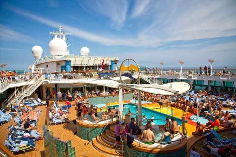 Crowded cruise ship pool deck with passengers relaxing, swimming, and enjoying activities under a sunny sky.
