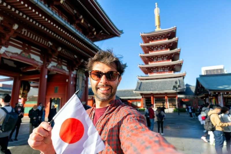 young tourist enjoying summer holiday in Tokyo, Japan - Traveling life style concept with smiling man taking selfie on city street with japan flag- Tourism and summertime vacation concept