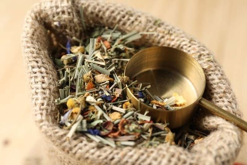 Close-up of dried herbal tea blend with a metal scoop inside a burlap sack, featuring mixed leaves, flowers, and botanical ingredients.