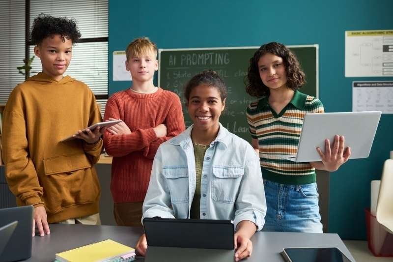 Group of students using tablets and laptops in a classroom focused on AI prompting and technology learning.