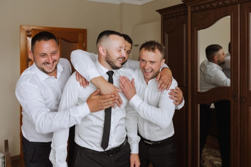 Stylish groom laughing and having fun with groomsmen while getting ready in the morning for wedding ceremony
