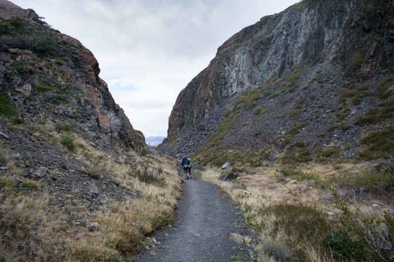 Lone hiker trekking through a narrow rocky canyon on a wilderness trail, surrounded by steep cliffs and rugged terrain.