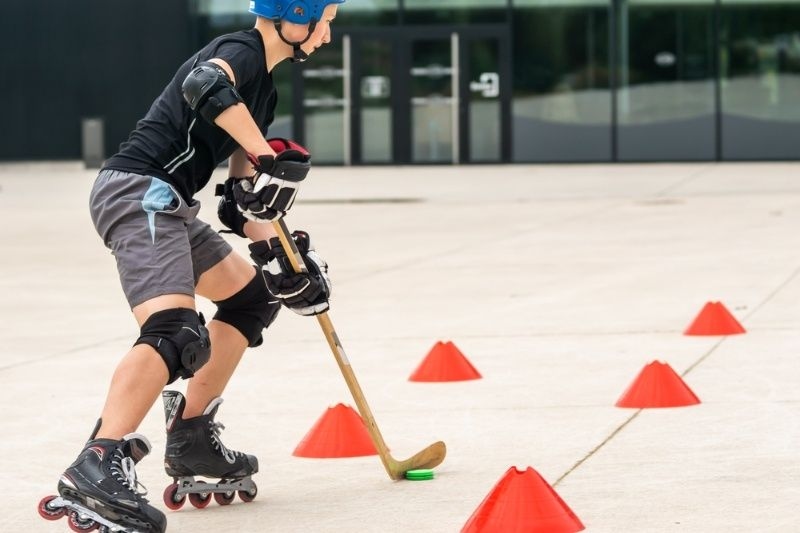 Person wearing protective gear practicing roller hockey, skating through cones while controlling a puck with a stick.