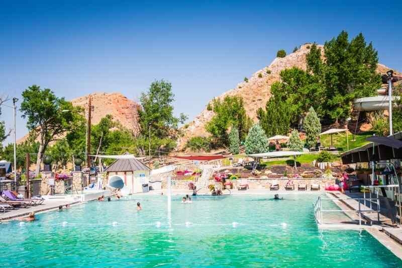 Outdoor hot springs pool with people relaxing, surrounded by trees and rocky hills under a clear sky.
