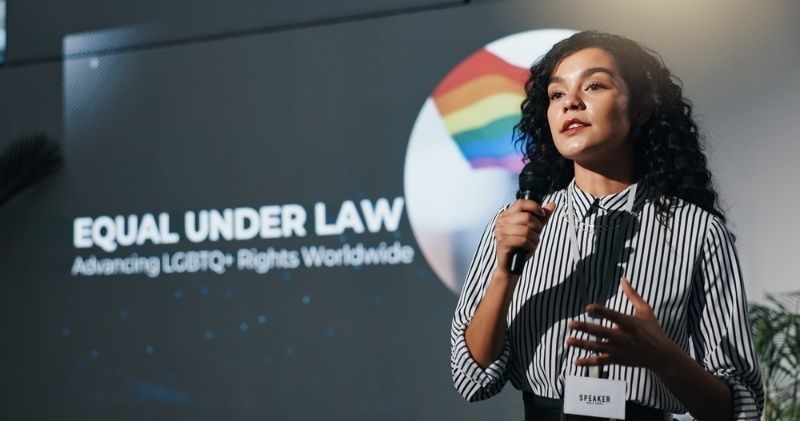 Speaker presenting on stage with text about LGBTQ+ rights and equality in the background.