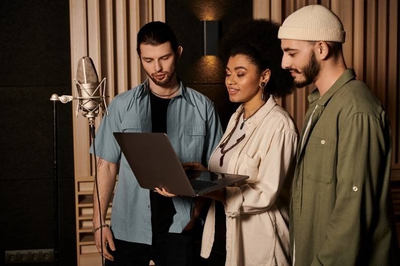 3. Three musicians standing in a recording studio reviewing content on a laptop beside a professional microphone.