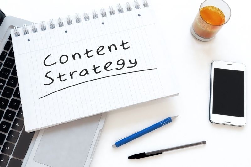 Notebook labeled ‘Content Strategy’ on a desk with laptop, smartphone, pen, and a glass of juice.