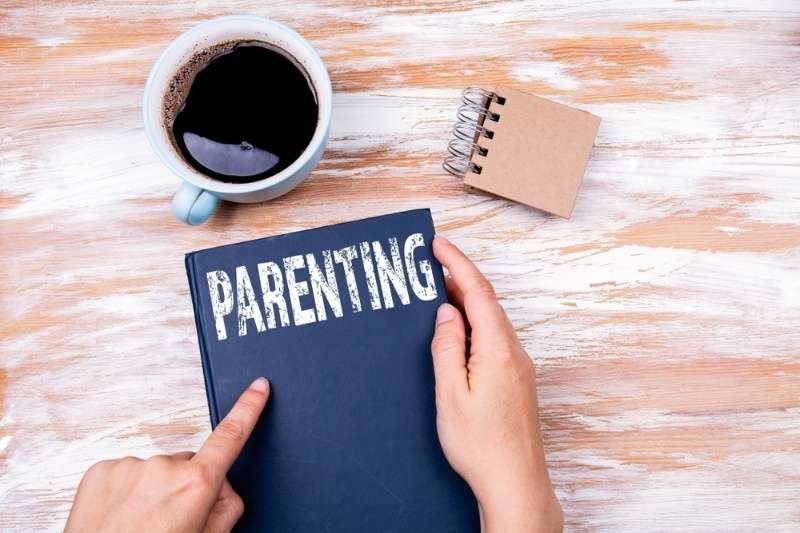 Hands holding a book titled “Parenting” on a desk beside a cup of coffee and a small notebook.