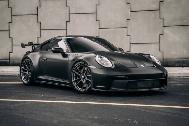 Matte black Porsche 911 parked on a city street beside a modern concrete wall.
