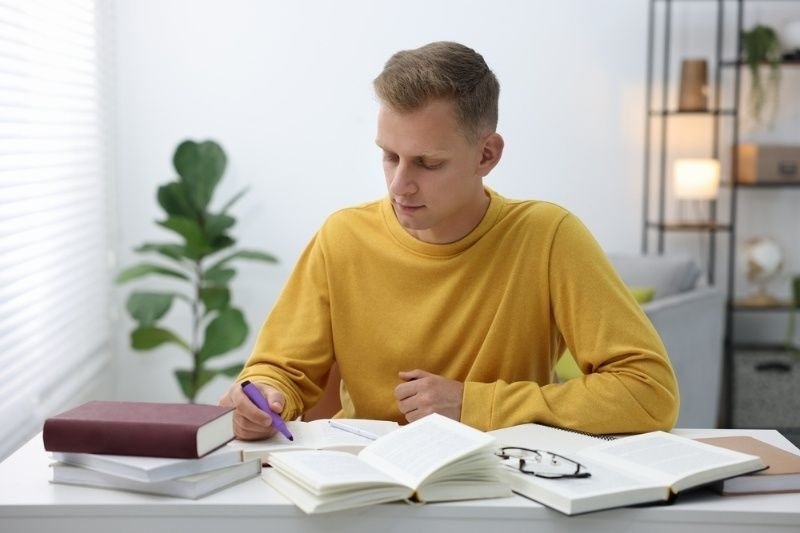 Student studying at a desk with open books and notes, preparing for exams in a bright indoor setting.