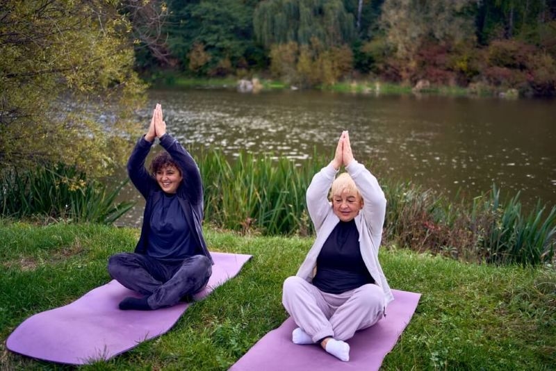 A middle-aged woman teaches her old mother somatic exercises and yoga to maintain health by the river in autumn