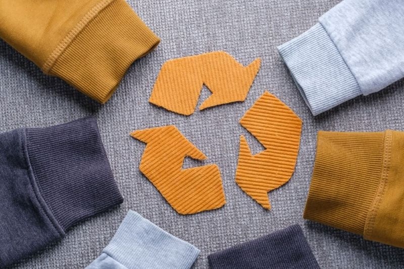 Sweater sleeves in shades of gray, mustard, and light blue surround a recycling symbol made from orange fabric