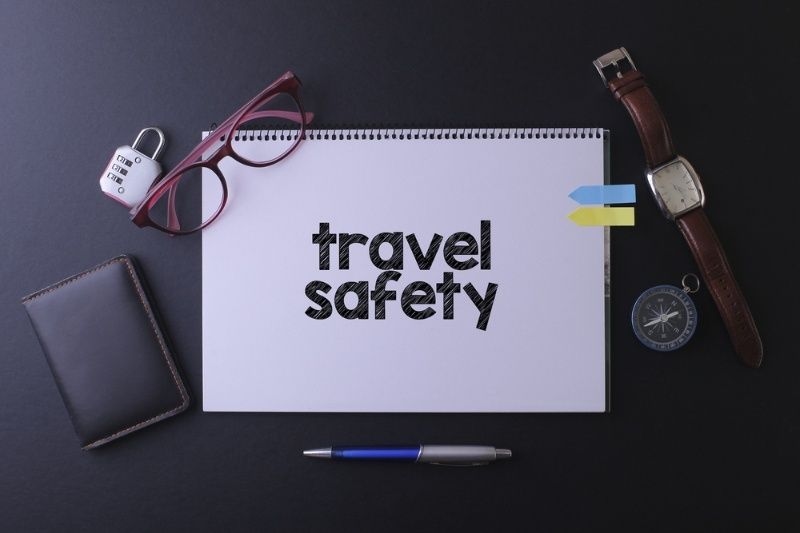 Notebook labeled ‘travel safety’ surrounded by essentials like glasses, wallet, lock, watch, and compass on a dark surface.