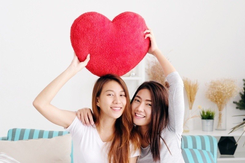 Two smiling people sitting on a couch indoors, holding a large heart-shaped pillow above their heads.