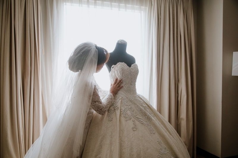 Bride examining an elegant lace wedding dress near a bright window.