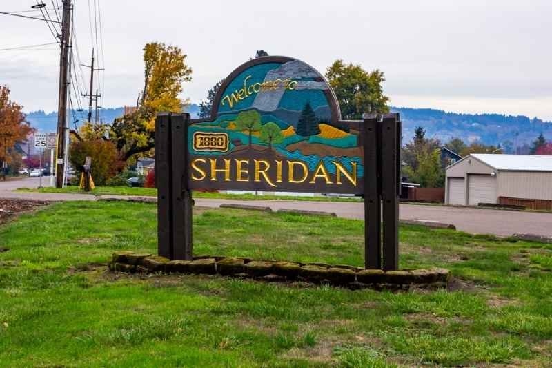 Welcome sign for Sheridan displayed at the town entrance, surrounded by grass, roadside, and residential buildings.