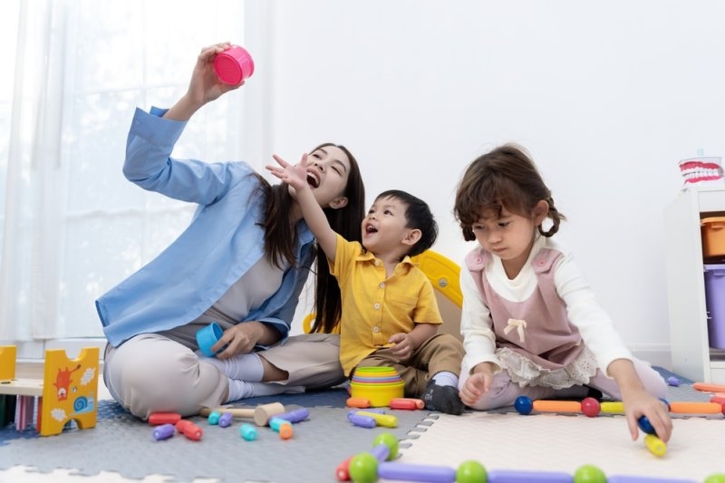 A teacher playing with two young children in a classroom, using colorful toys and stacking games to encourage learning through play.