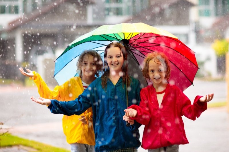 Three children wearing colorful raincoats smile beneath a rainbow umbrella while enjoying the rain outdoors.