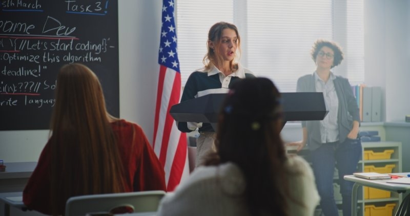 American Female Student Delivers Speech With US Flag in Front of Classmates Sitting Desks. Concept Civic Education, Public Speaking Skills, and Student Voice in American Education System. Dolly Shot.