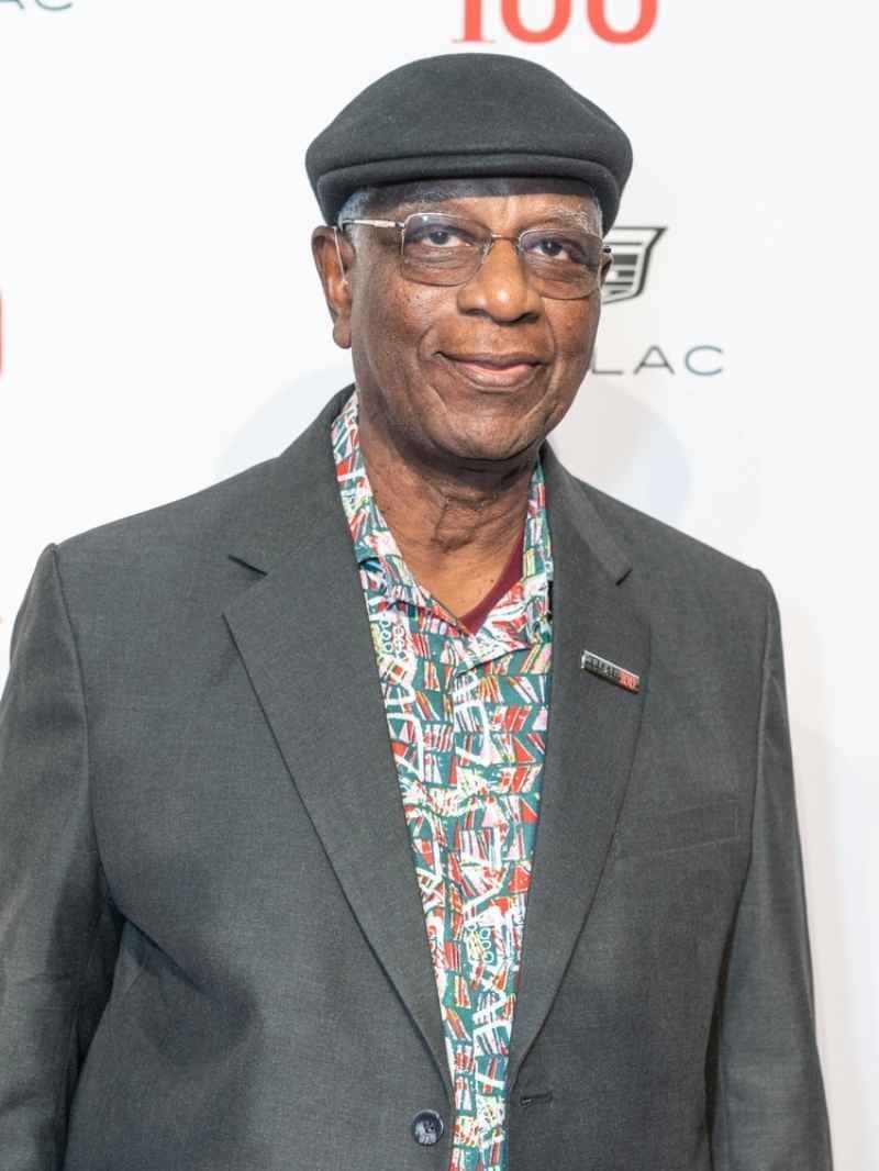 A professionally dressed older man wearing glasses and a flat cap poses for a photograph at a formal event.
