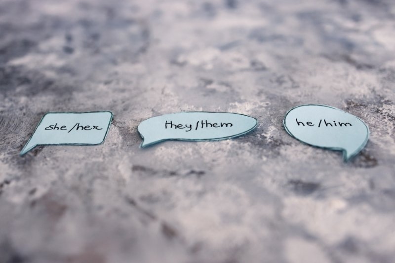 Speech bubbles displaying “she/her,” “they/them,” and “he/him” pronouns on a textured surface.
