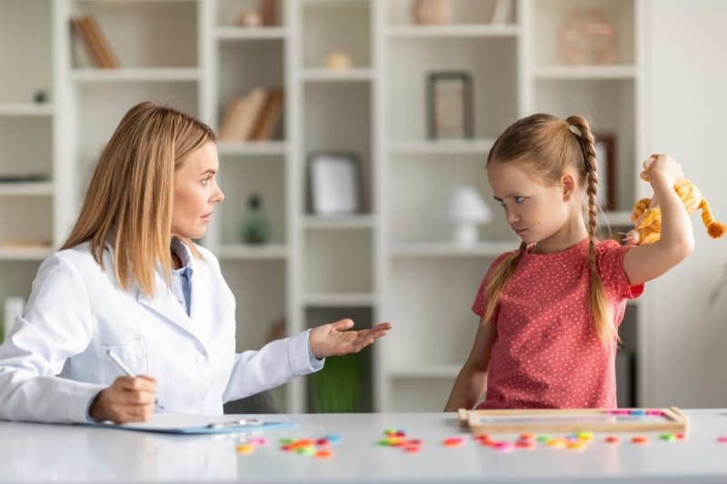 Healthcare professional observing a young girl holding a toy while seated at a table during a behavioral session.