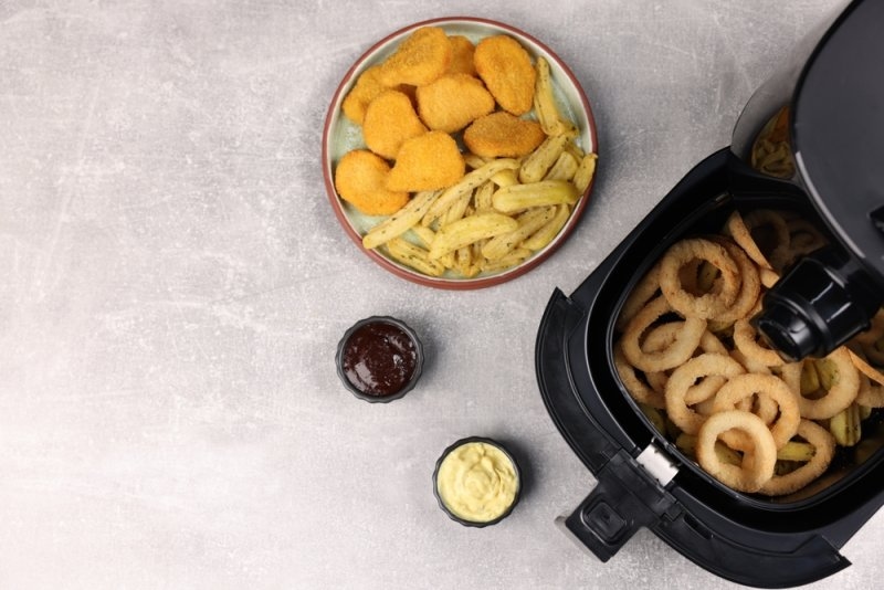 Air fryer with tasty squid rings, chicken nuggets, french fries and sauces on grey table, flat lay. 