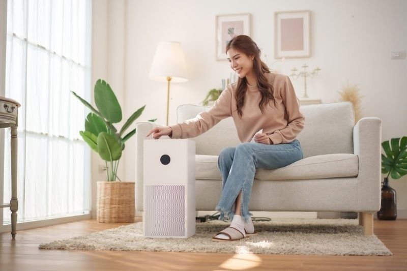Woman sitting on a living room sofa while turning on a modern air purifier in a bright, comfortable home environment.