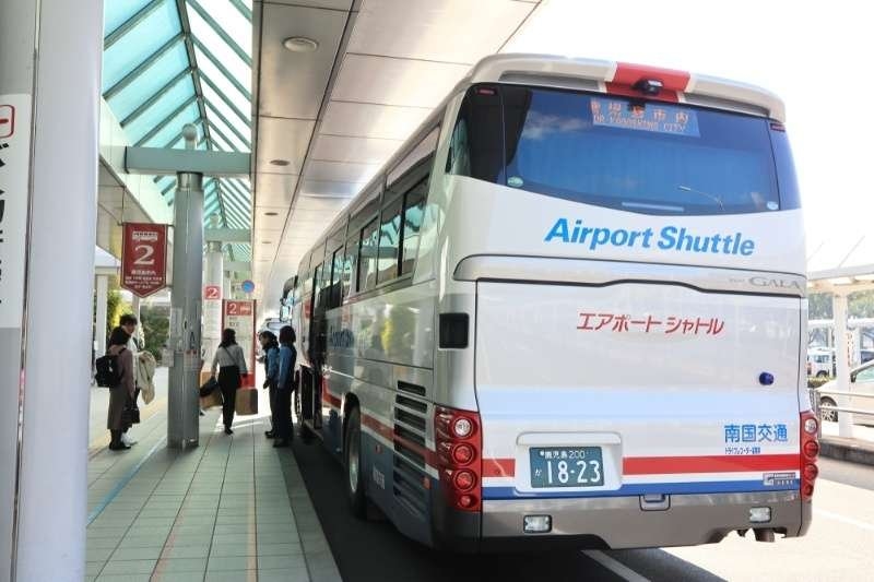 Airport shuttle bus stopped at a passenger pickup area outside a terminal, with travelers boarding and waiting nearby.