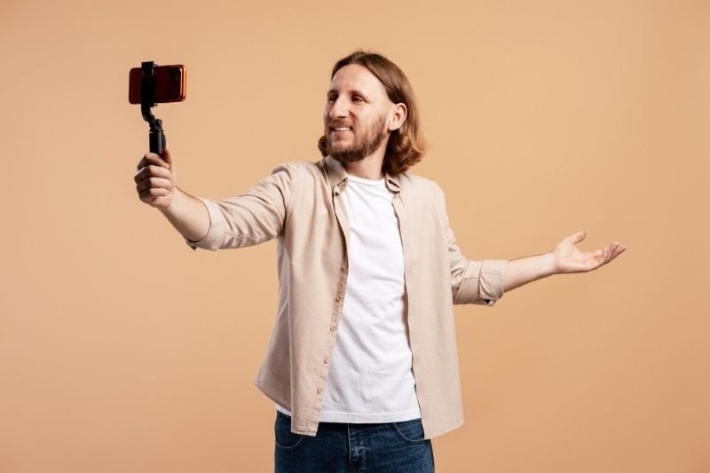 A smiling man holds a smartphone mounted on a handheld gimbal or stabilizer, appearing to record himself against a plain peach-colored background.